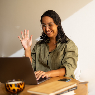 Woman in green shirt waving at a laptop with a notebook and phone on the table, starting an online consult to refine her office wellbeing workplace set up